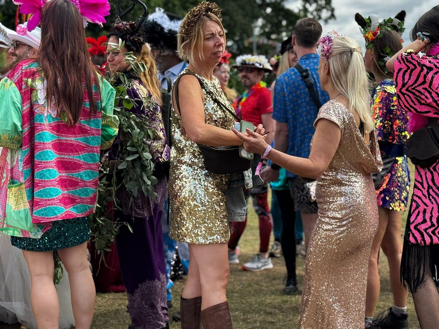 People dressed in bright, sequinned and floral festival outfits standing together outdoors. Two women in gold and rose‑gold dresses are holding hands in the foreground, surrounded by others in colourful patterned clothing. It is a lively, creative festival scene.