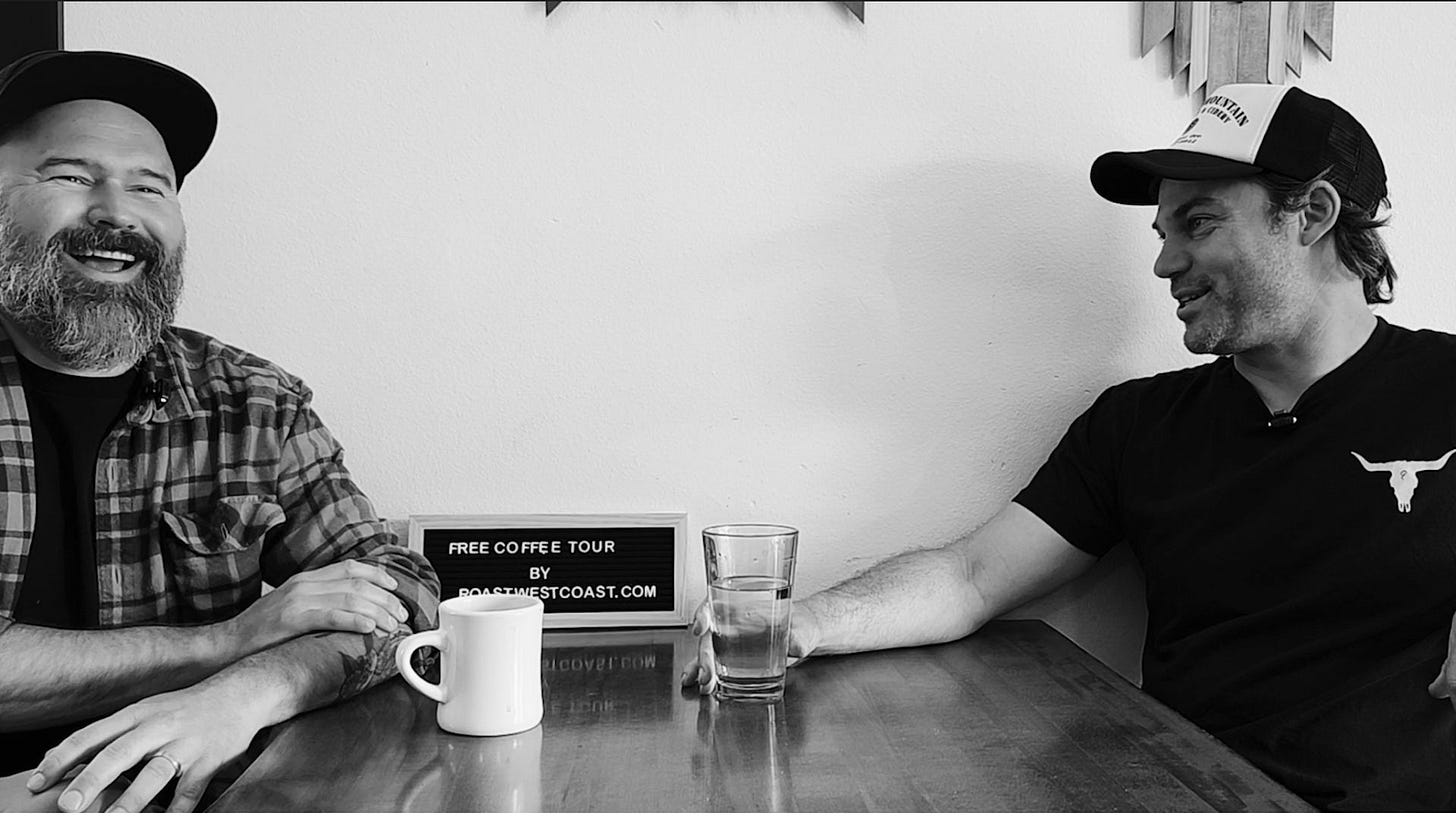 A black and white photo of two men sitting at a table in a coffee shop with a white wall behind them. The man on the left has a big beard and is laughing emphatically. The man on the right is talking.