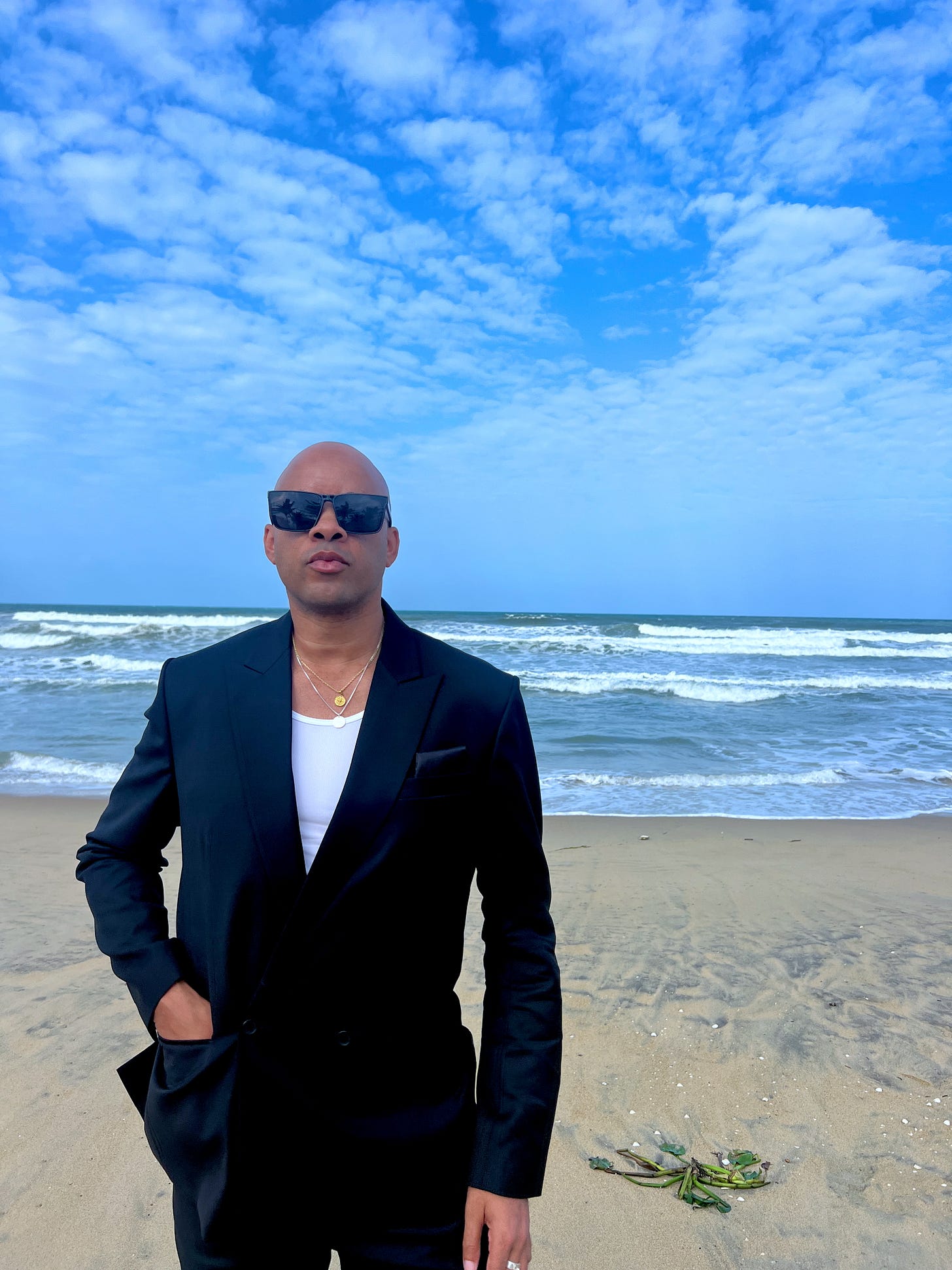 Ethan Ward wearing a black suit and sunglasses stands on the beach with the sea and blue sky behind him. Ethan Ward wearing a black suit and sunglasses stands on the beach with the sea and blue sky behind him.