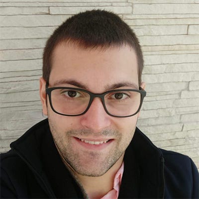 Homem de óculos e cabelos curtos, usando casaco preto e camisa vermelha, tendo ao fundo uma parede de pedra