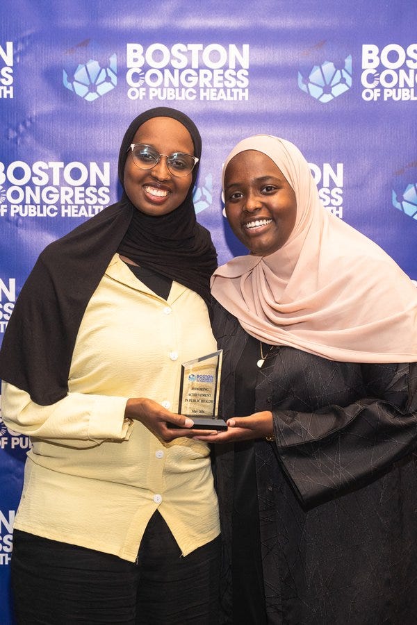 Sirad Hassan, MS, in a yellow blouse and black hijab, beside a colleague in a pink hijab, with her 40 Under 40 statue on the BCPH backdrop.