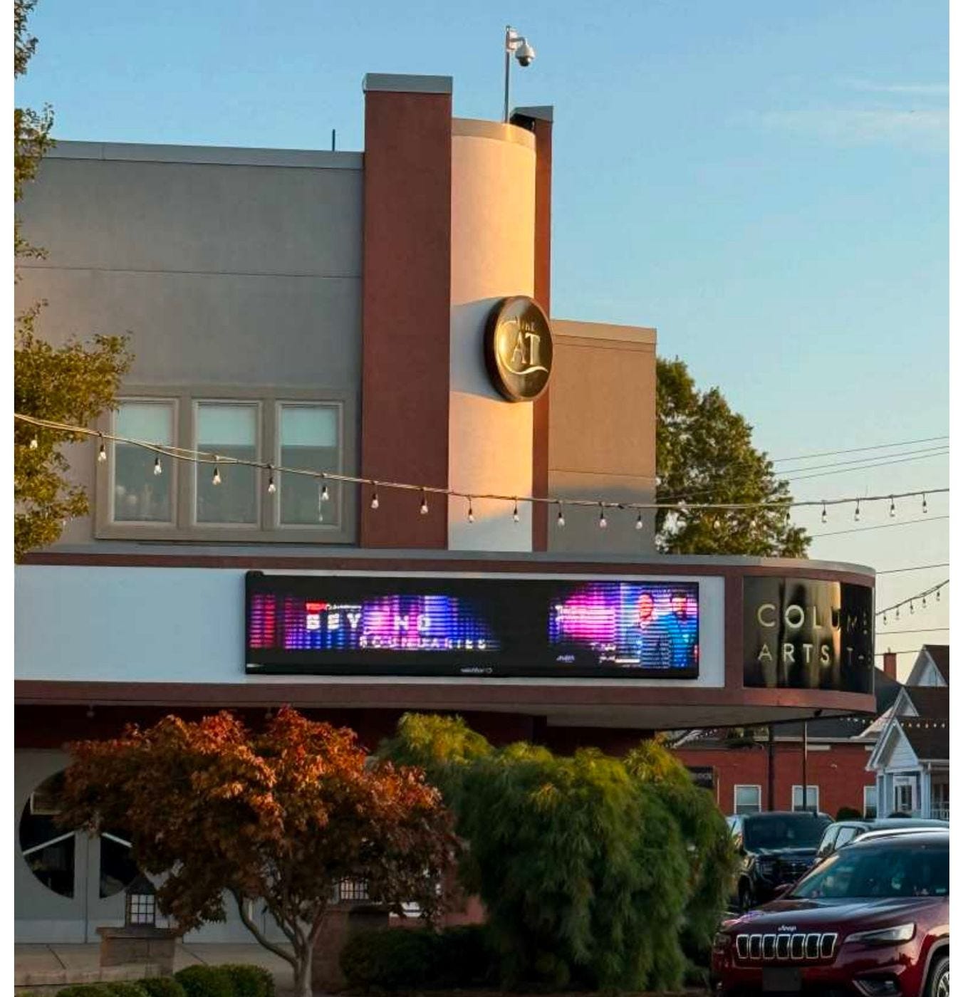 The Columbiana Arts Theater at golden hour. A gold roundel reading the CAT sits above a lit LED marquee running TEDxColumbiana Beyond Boundaries, with string lights strung overhead and a house next door. The Columbiana Arts Theater at golden hour. A gold roundel reading the CAT sits above a lit LED marquee running TEDxColumbiana Beyond Boundaries, with string lights strung overhead and a house next door.