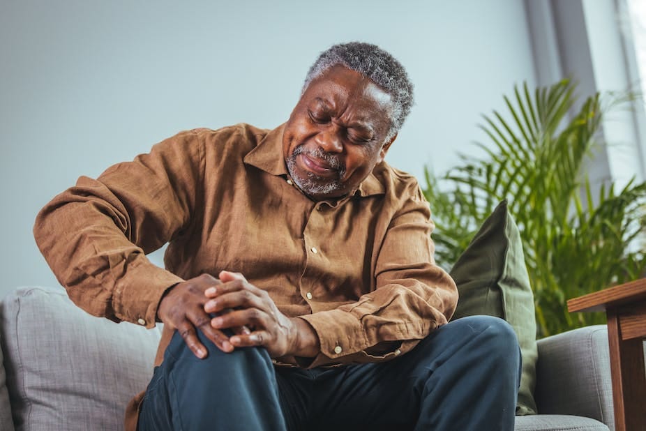 An older man of African ethnicity sits on a couch holding his knee, grimacing in pain. He is wearing dark blue trousers and a camel-coloured shirt An older man of African ethnicity sits on a couch holding his knee, grimacing in pain. He is wearing dark blue trousers and a camel-coloured shirt