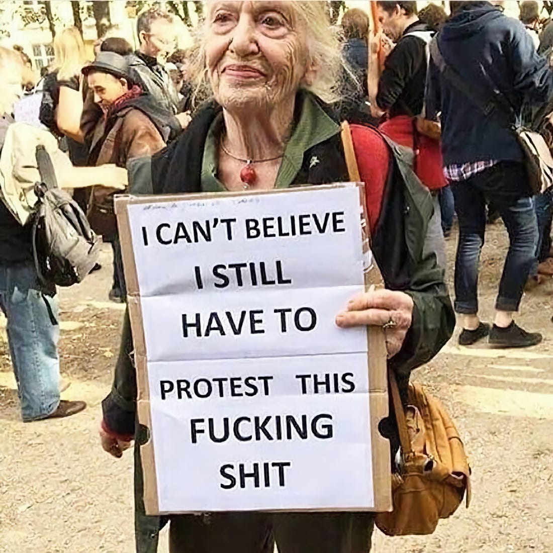 Elderly woman at a protest holding a handmade sign: I can't believe I still have to protest this fucking shit