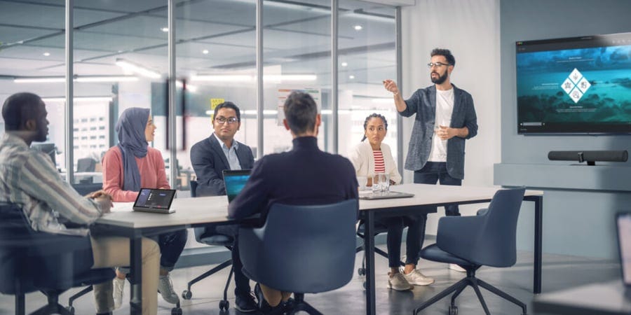 A group of five people are sitting around a conference table listening to a man giving a presentation while pointing at a screen displaying a graphic. A group of five people are sitting around a conference table listening to a man giving a presentation while pointing at a screen displaying a graphic.
