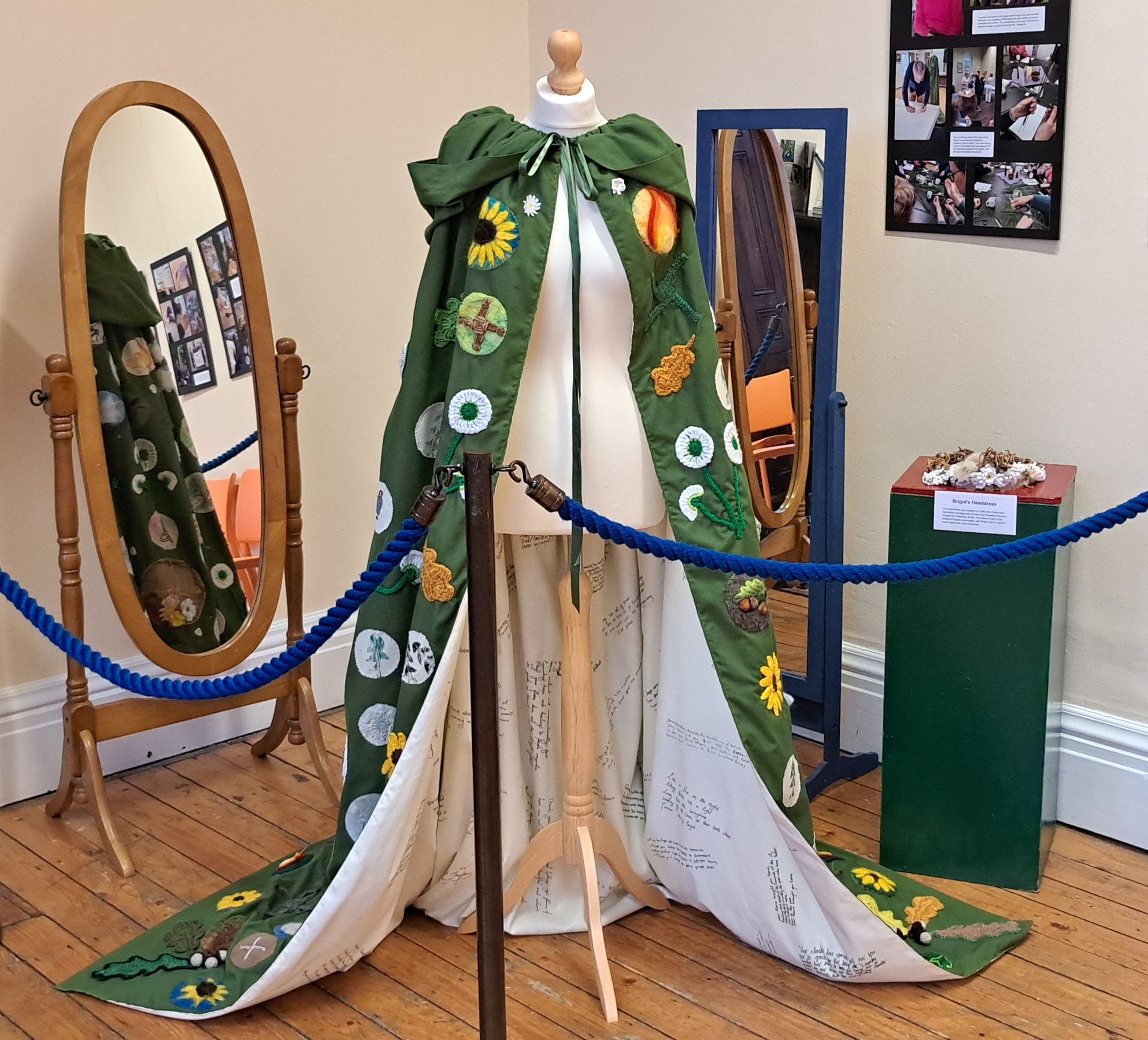 The green decorated cloak on a dressmakers dummy on display in the museum. Behind it are two oval mirrors so viewers can see the details on the back of the cloak. The cloak has been pulled and draped at the front so viewers can see and read some of the messages written into the lining, which is cream in colour.