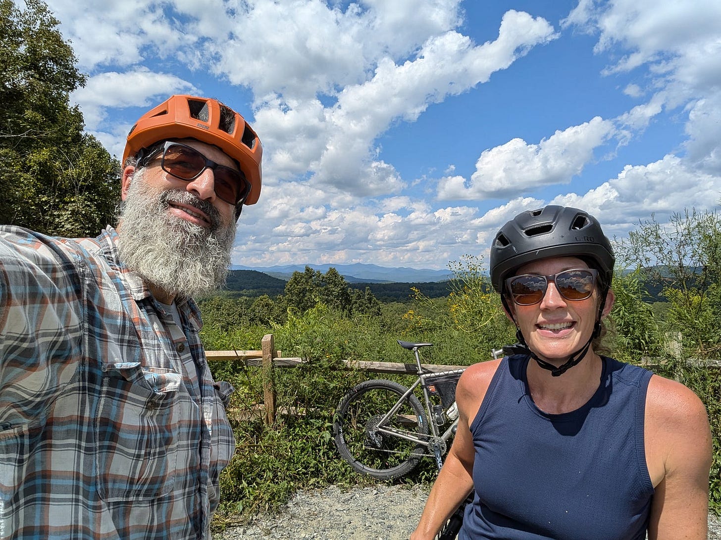 The author and his wife, both wearing bike helmets and sunglasses, smile in front of a scenic overlook in DuPont State Forest. A gravel bike leans against a wooden fence behind them. The sky is bright blue with scattered white clouds above a lush, green mountain landscape. The author and his wife, both wearing bike helmets and sunglasses, smile in front of a scenic overlook in DuPont State Forest. A gravel bike leans against a wooden fence behind them. The sky is bright blue with scattered white clouds above a lush, green mountain landscape.
