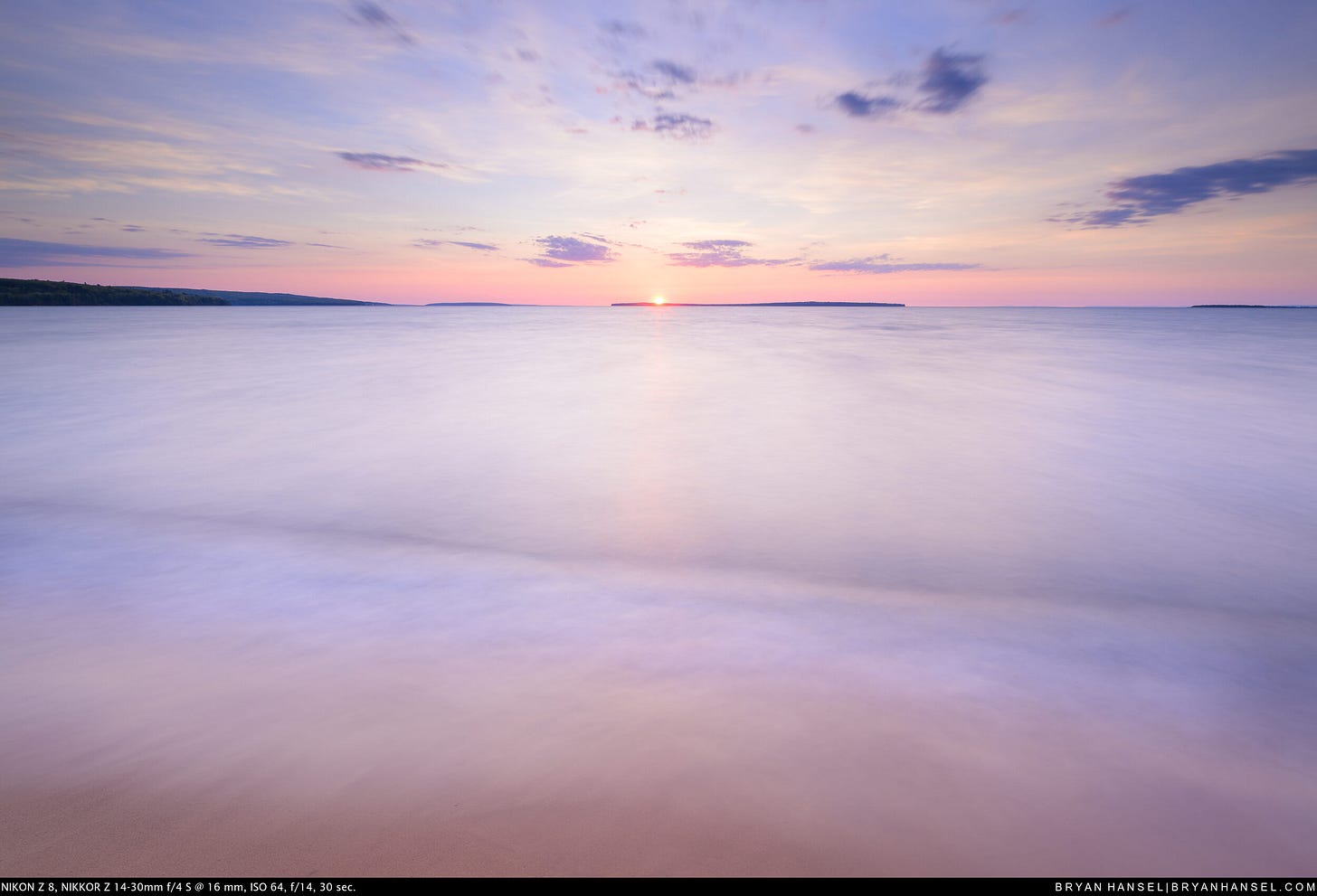 The sun sets precisely on the horizon between two distant islands on Lake Superior, its warm orange glow reflecting in silky 30-second exposed water washing over a sandy beach under a wide, softly clouded sky.