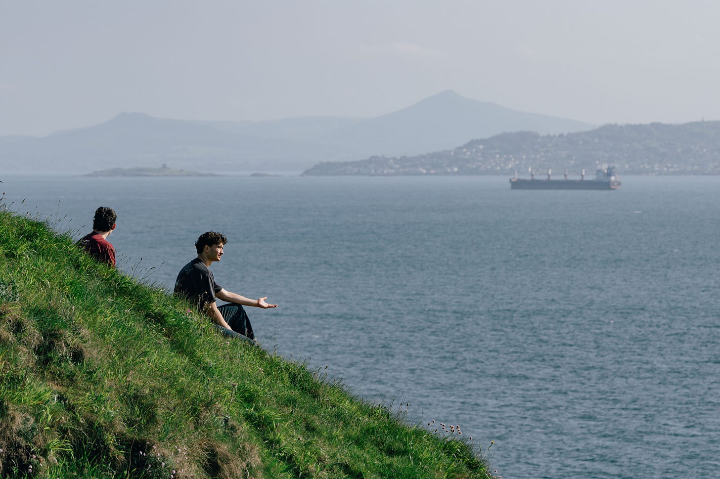 Two men sit on the green hillside above the sea, one gesturing mid-conversation, with Dublin Bay and a cargo ship in the background. Two men sit on the green hillside above the sea, one gesturing mid-conversation, with Dublin Bay and a cargo ship in the background.