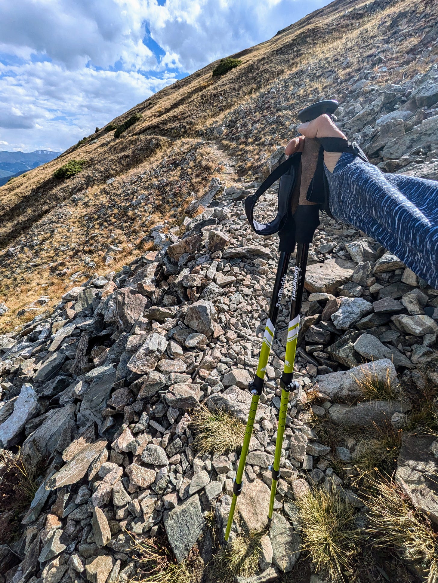 Hand holding trekking poles on a rocky hillside trail, with open alpine terrain and clouds in the distance. Hand holding trekking poles on a rocky hillside trail, with open alpine terrain and clouds in the distance.