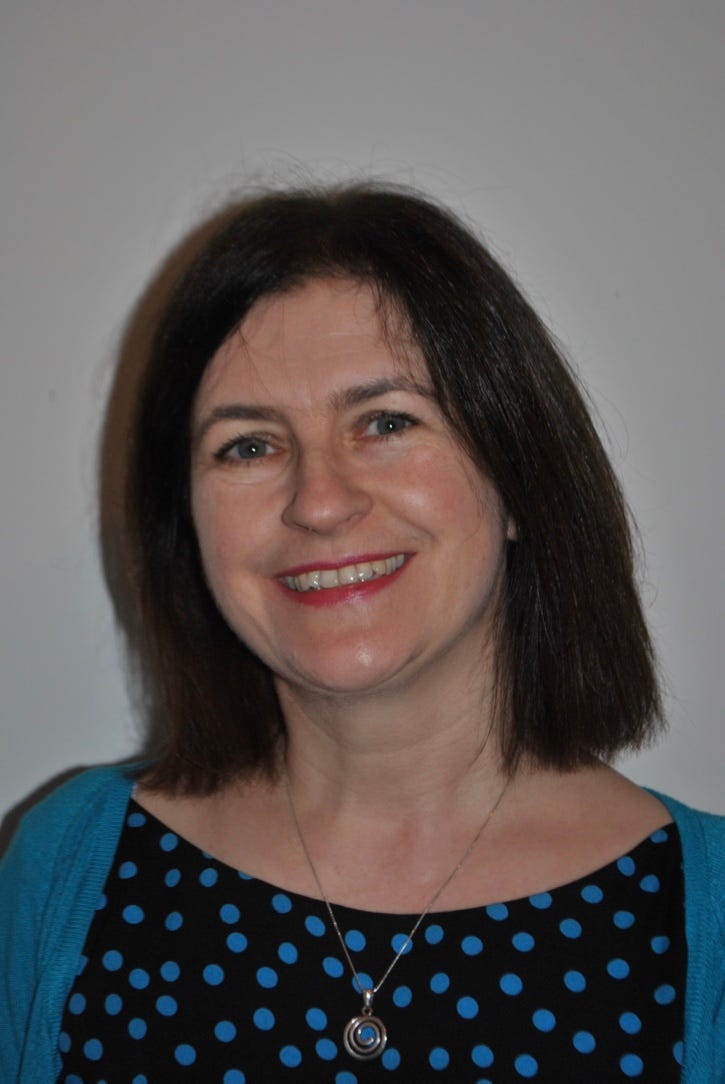 A photo of Sheena. She has brown hair to her shoulders, a big smile, and is wearing a black dress with blue spots and a necklace.
