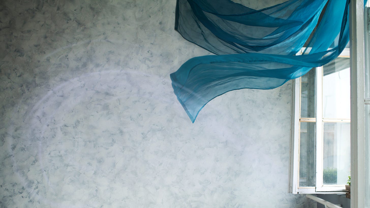 A imagem mostra um ambiente interno tranquilo e iluminado. Há uma parede com textura marmorizada em tons de cinza e branco, que dá um toque artístico e sofisticado ao espaço. Uma janela está aberta, permitindo que a luz natural entre suavemente no cômodo. Perto dela, um tecido azul translúcido está pendurado, esvoaçando levemente com a brisa que entra. Essa combinação cria uma atmosfera serena, quase poética — como se o tempo estivesse passando devagar ali.