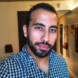 Jovem de porte atlético, cabelo curto, escuro e cacheado, olhos castanhos-escuros e uma barba escura. Sua expressão facial é neutra e serena, e ele olha diretamente para a câmera. Veste uma camisa clássica de botões com estampa xadrez em preto e branco. Jovem de porte atlético, cabelo curto, escuro e cacheado, olhos castanhos-escuros e uma barba escura. Sua expressão facial é neutra e serena, e ele olha diretamente para a câmera. Veste uma camisa clássica de botões com estampa xadrez em preto e branco.
