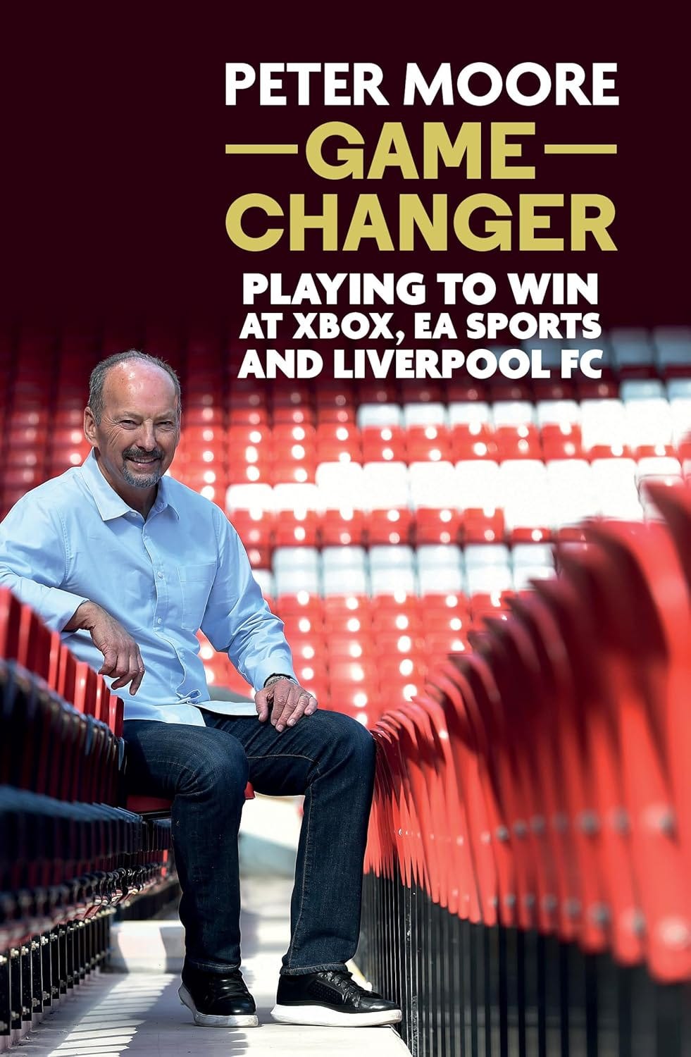 Book cover for Game Changer by Peter Moore, featuring the author seated in a stadium's red seats, wearing a light blue button-up shirt and jeans. The bold text above him reads “PETER MOORE — GAME CHANGER — Playing to Win at Xbox, EA Sports and Liverpool FC,” highlighting his leadership roles across gaming and sports industries. The setting and layout emphasize his connection to competitive environments and major entertainment brands.