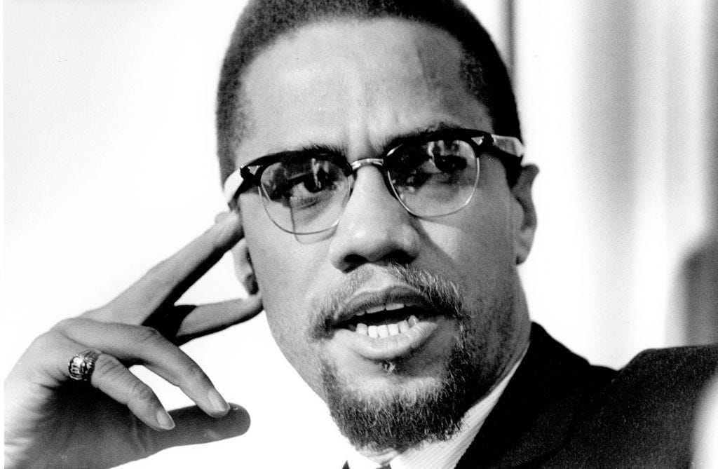 Photo of Malcolm X in mid-sentence, with a finger pressed to his temple, looking off to the left of the frame.