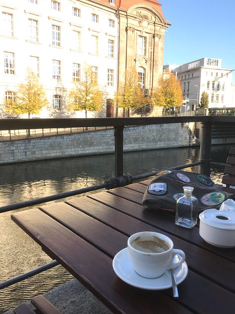Views of a coffee shop, cats staring at a front-loading German washing machine, a woman in a large rain poncho, a cat sleeping on an induction stove burner, a courtyard filled with flowers, and elaborate stonework facade in Berlin, coffee on a table next to a river, a dutch style bike parked next to a tree with yellow leaves covering the ground and a man in a sofa with three cats cuddled up with him