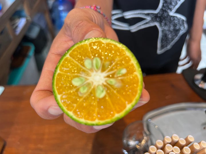 A man at a counter pressing a halved orange on a hand press, the juice running into a paper cup. An orange sliced in half, the skin green all the way around, the flesh ripe and orange inside. 