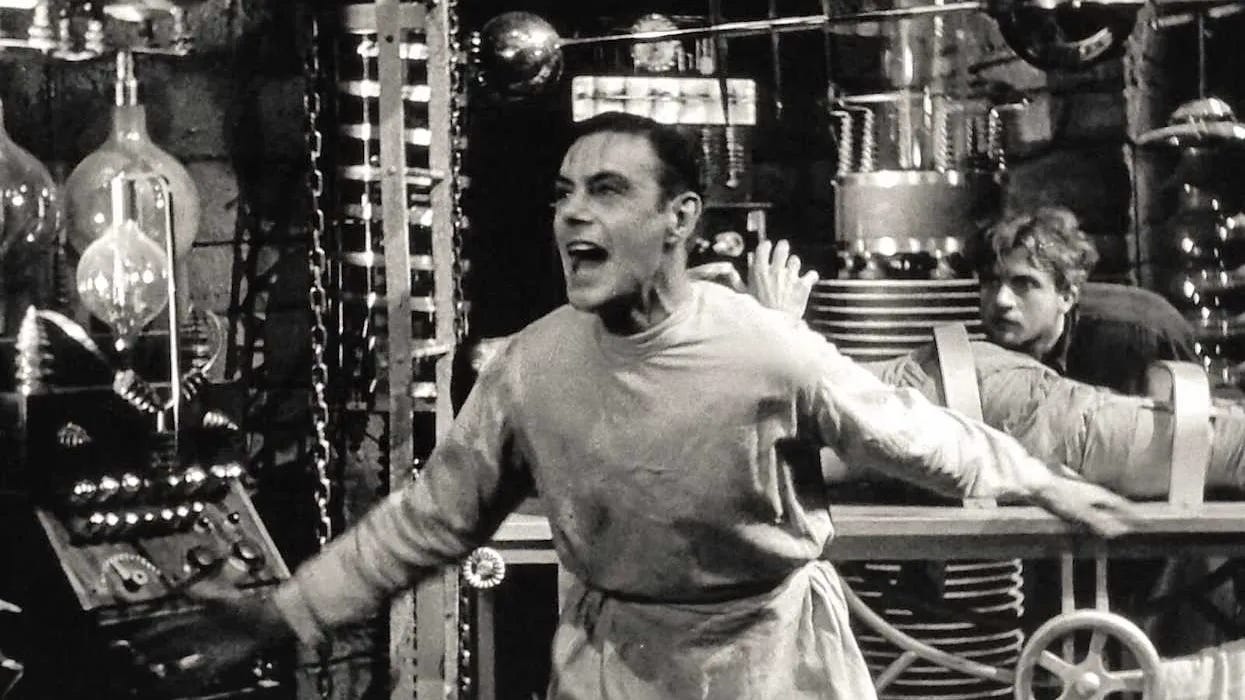 Black-and-white film still showing a man in a laboratory shouting excitedly beside electrical equipment, glass apparatus and machinery, while another man lies strapped to a table behind him. Black-and-white film still showing a man in a laboratory shouting excitedly beside electrical equipment, glass apparatus and machinery, while another man lies strapped to a table behind him.