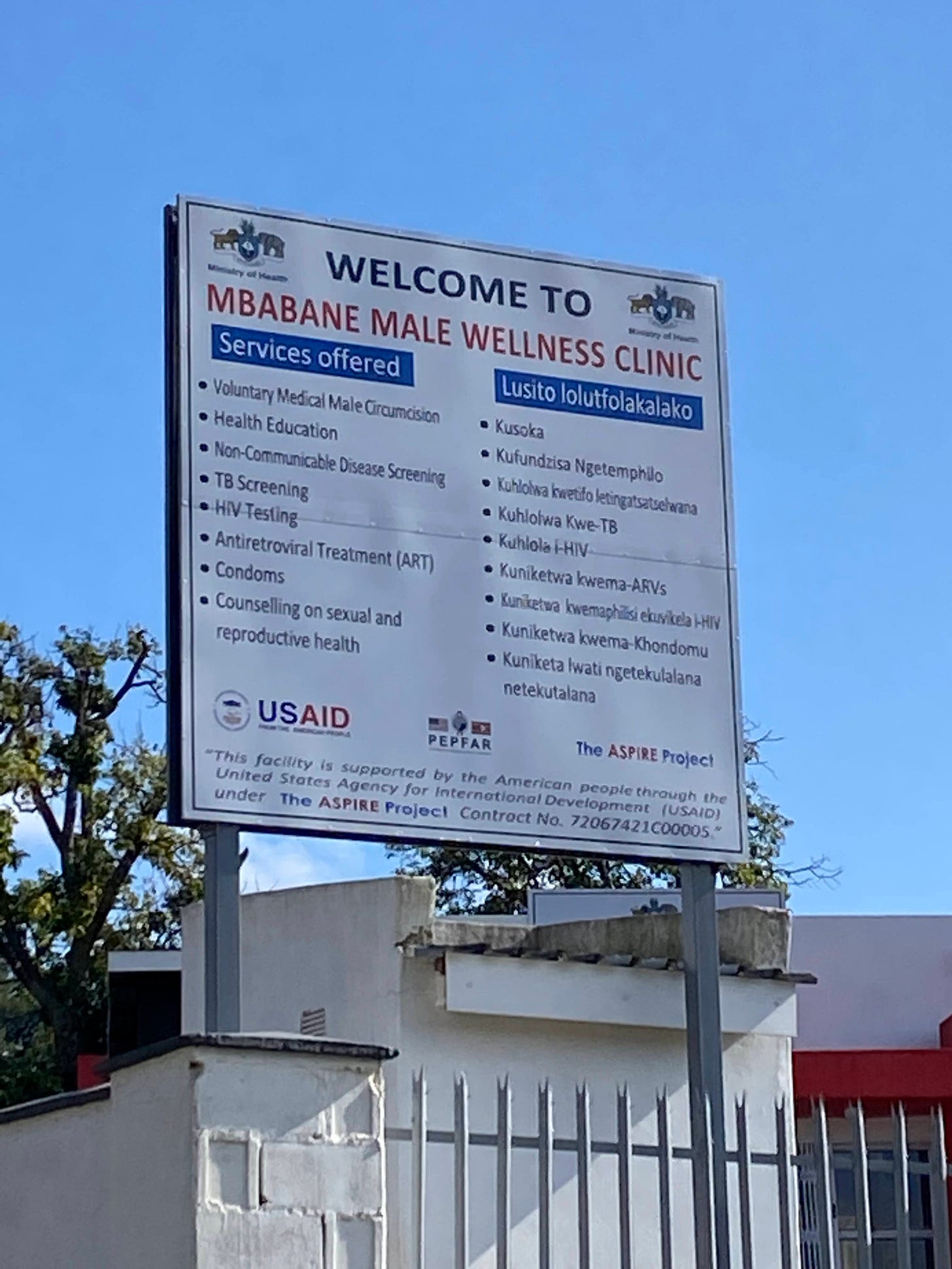 A sign in front of a clinic reads in English and Siswati, “Welcome to Mbabane Male Wellness Clinic,” with a list of services offered, the majority being for sexual and reproductive health. Underneath it are the logos of USAID, PEPFAR, and the ASPIRE project, and the grant number. 