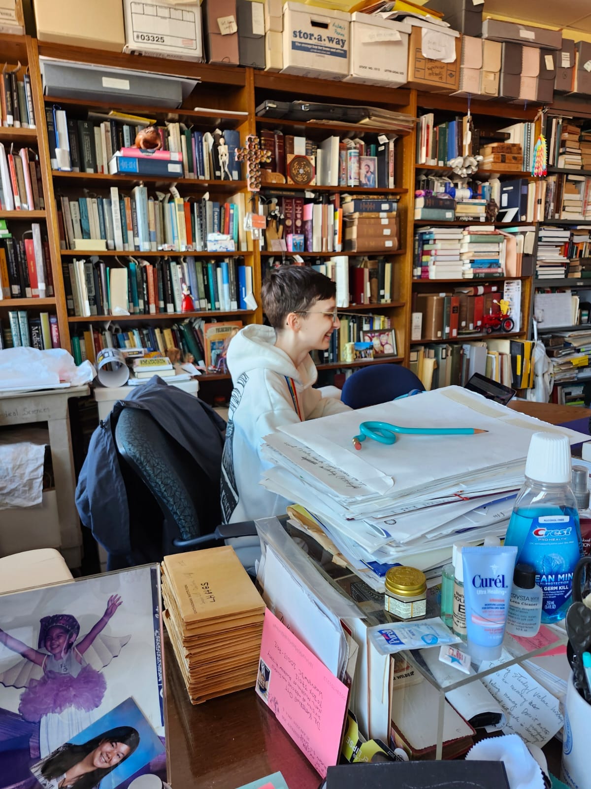 Beck sitting at a desk in a cluttered office filled with books, boxes and papers. Beck is smiling, happily looking at their laptop.