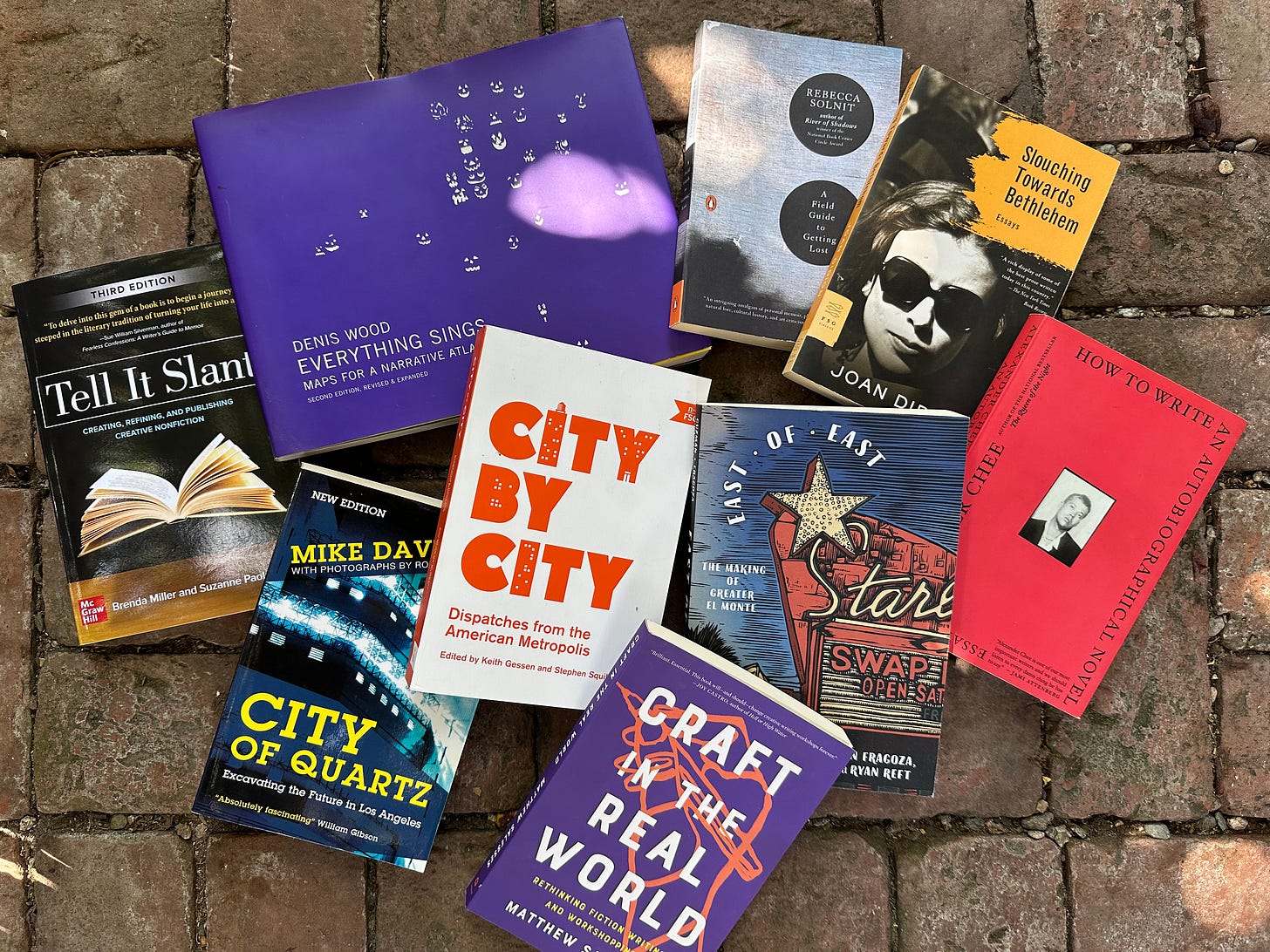Photo of a mess of books on a brick walkway. Books includeL City of Quarts by Mike Davis, Tell it Slant by Brenda Miller and Suzanne Paola, Everything Sings by Denis Wood, Craft in the Real World by Matthew Salesses, City by City (edited collection), East of East (edited collection), How to Write an Autobiographical Novel by Alexander Chee, A Field Guide to Getting Lost by Rebecca Solnit, and Slouching Towards Bethlehem by Joan Didion