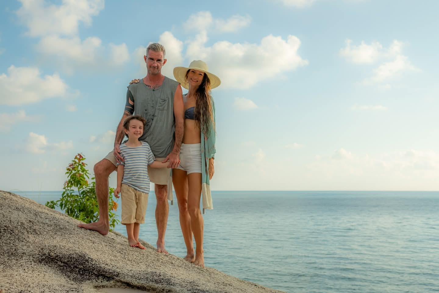 A smiling husband and wife stand on a rock by the ocean with their adorable young son.