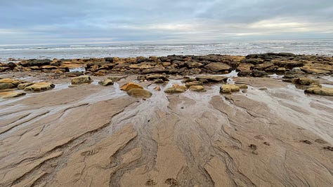 Six different shots of the beach with sand, rocks and shimmering water, several with blue skies, one with moody clouds and the sun peeking through.
