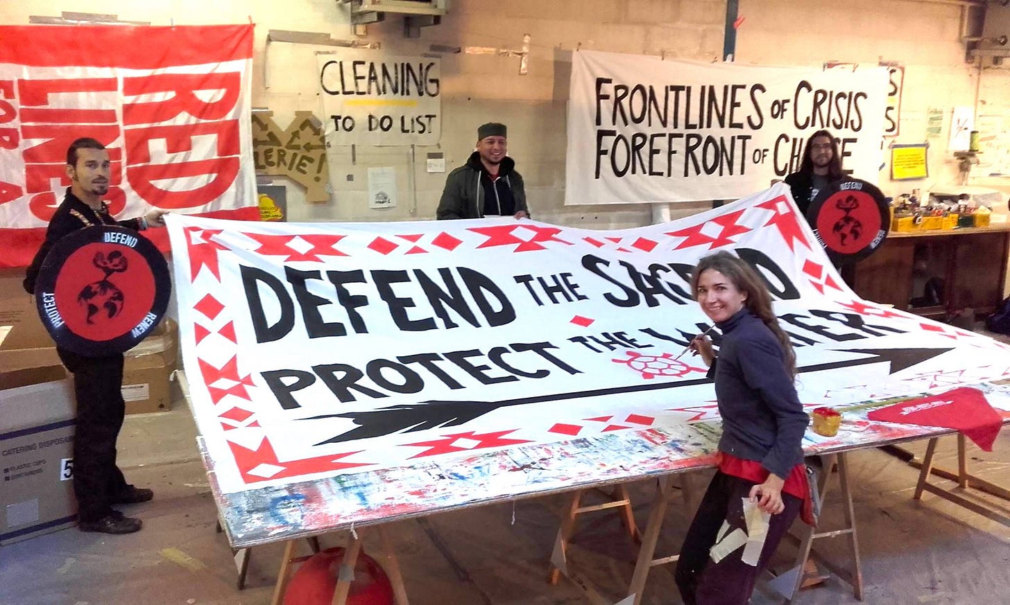Gera and three other activists paint banners for an upcoming action. The central banner--in red and black on a white background--reads "Defend the Sacred. Protect the Water." 