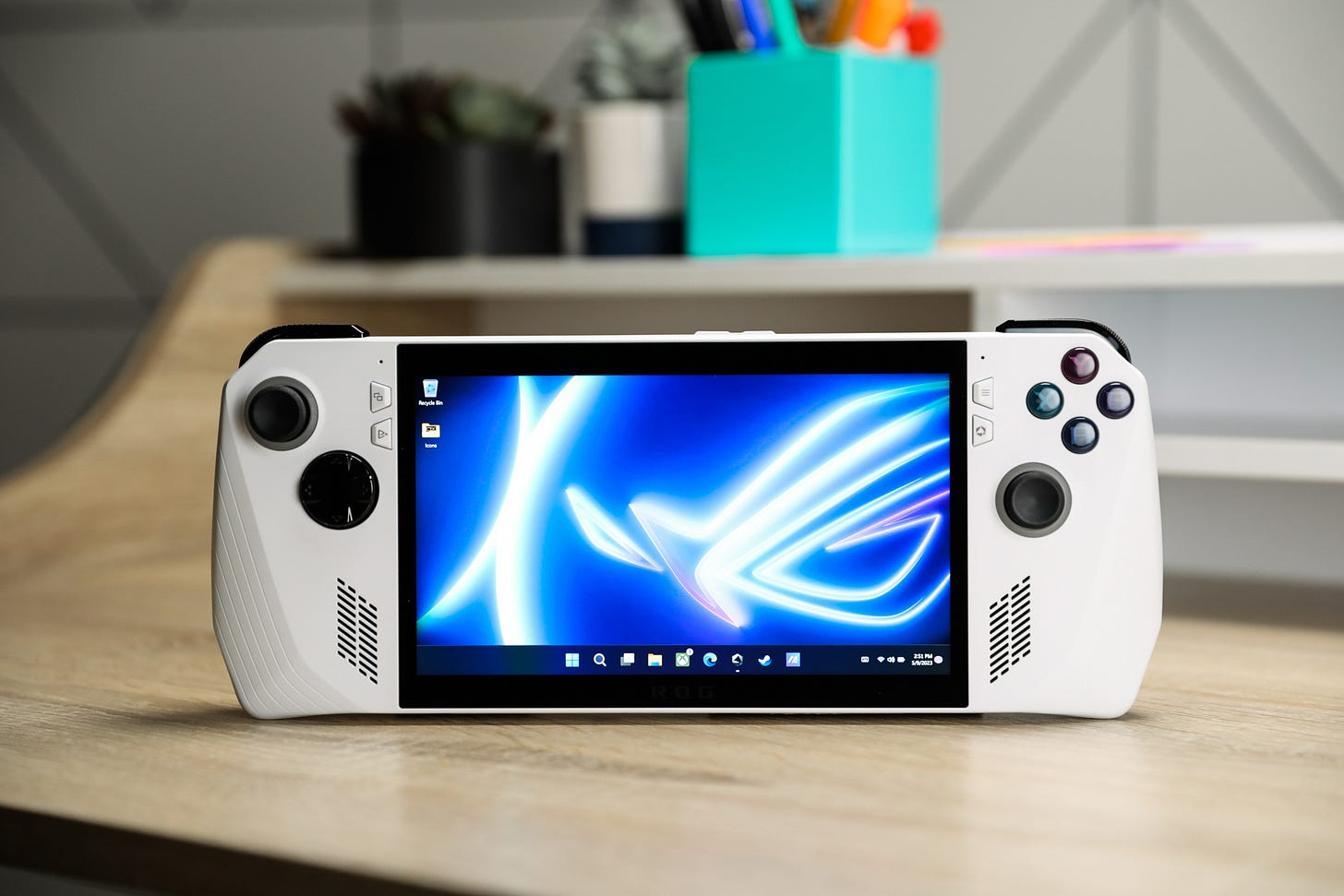 The Asus ROG Ally PC gaming handheld in white standing upright on a wood desk and showing a Windows homescreen.