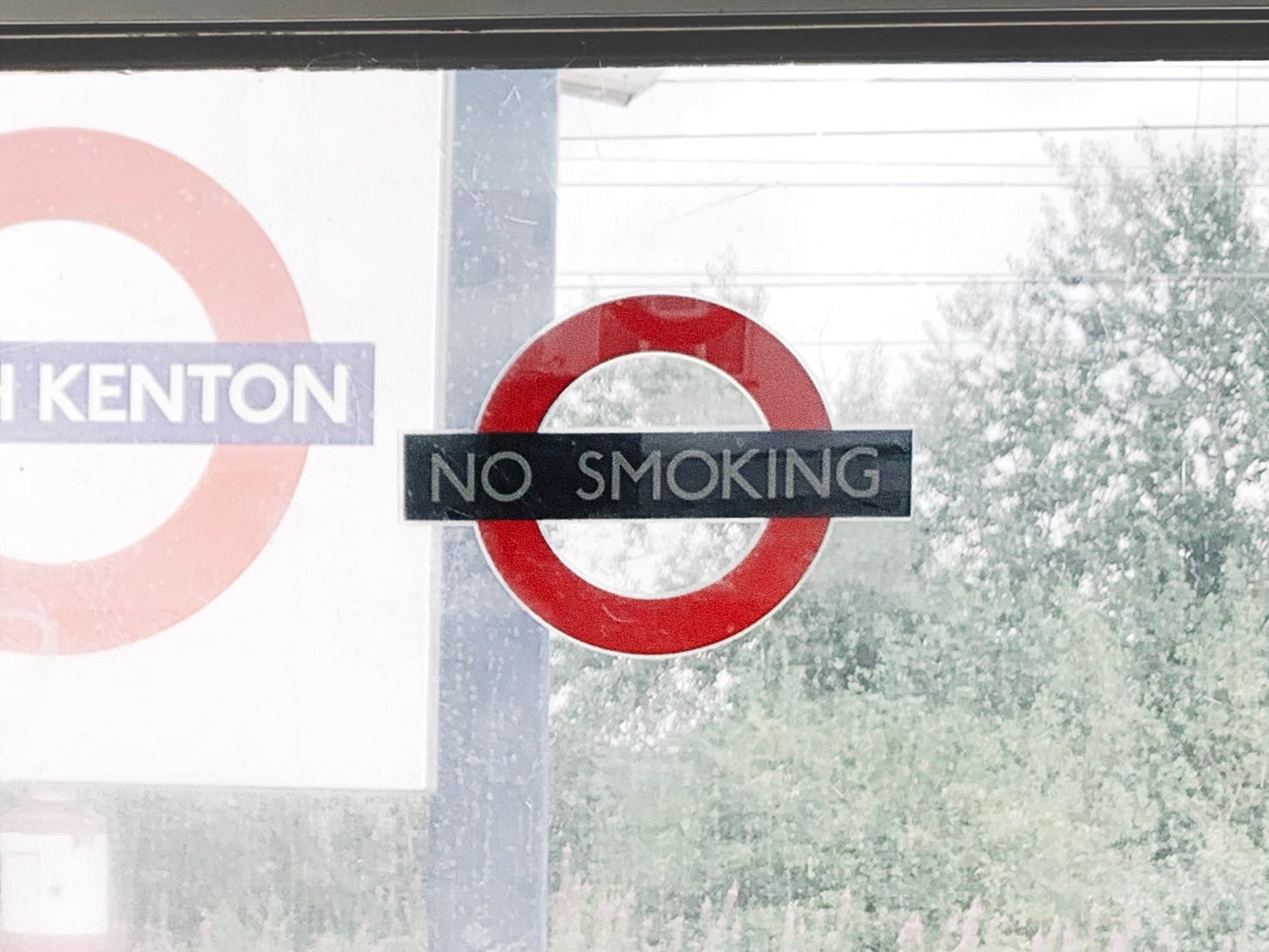 No Smoking Sign on a Window: The image features a "No Smoking" sign displayed on a window. The sign consists of a red circle with a horizontal black bar in the center, containing the text "NO SMOKING" in white capital letters. In the background, a partially visible sign reads "SOUTH KENTON," likely indicating a train or subway station. Additionally, there are trees and power lines visible, suggesting the setting is outdoors. The image emphasizes the importance of maintaining a smoke-free environment in public spaces.
