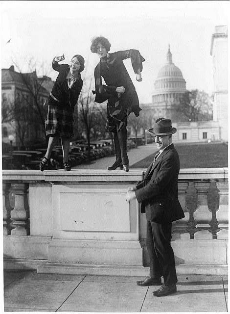 a black and white photo of two women dancing on top of a railing in Washington DC while a man in a suit stands on the ground next to them