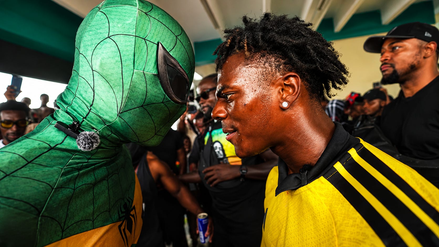 Enhanced cinematic image capturing a tense and unforgettable moment during a viral Jamaica livestream, as a masked figure and a young man in yellow stand face to face amid a packed crowd. The image has been recreated and enhanced for dramatic impact, with sharper detail, richer colour, and a more iconic landscape composition.