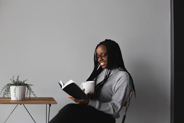 A woman with long black braids and glasses sits in a chair at a cafe table, holding a mug of coffee in one hand and an open book splayed in the other. On the table in front of her rests a plant in a small white pot.