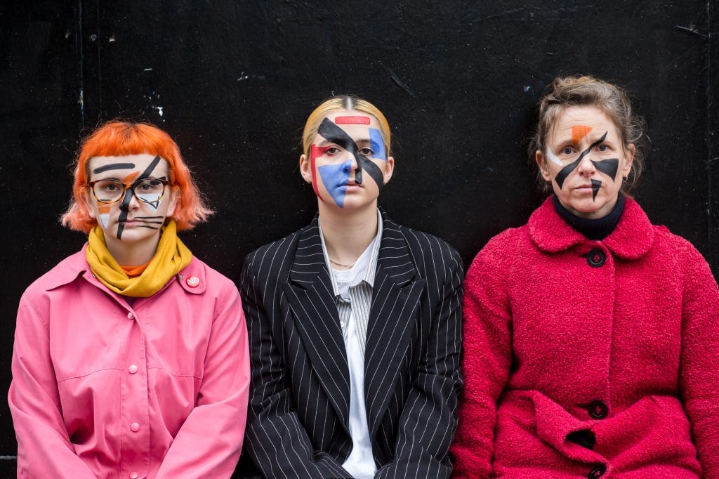 Emily, Evie and Anna sit in front of a black background facing the camera with straight faces. Each of them have bright blocky shapes painted on their faces in black, white, blue and red. Emily, Evie and Anna sit in front of a black background facing the camera with straight faces. Each of them have bright blocky shapes painted on their faces in black, white, blue and red.