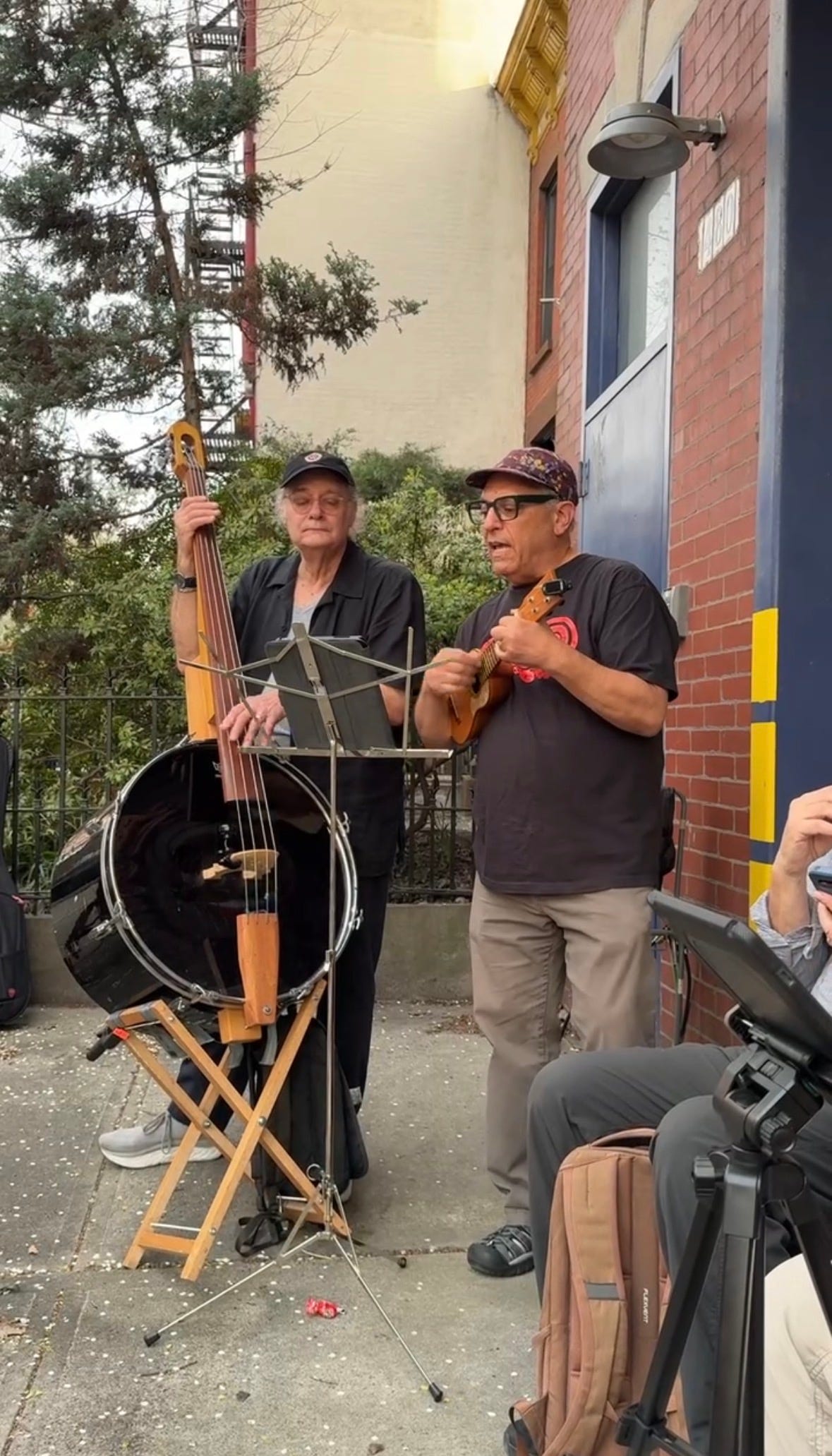 Two white men wearing hats and glasses stand on a sidewalk, one playing a stand-up bass with a body made of a drum, the other playing a ukelele Two white men wearing hats and glasses stand on a sidewalk, one playing a stand-up bass with a body made of a drum, the other playing a ukelele