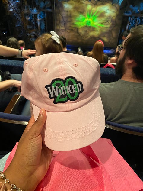 From top left to bottom right - going across horizontally. Image 1) Alexa is holding up a green frisbee that says Defying Gravity since 2003 in black font. The text is curved around the frisbee, in a half circle. In the center, there is a black stenciled picture of Elphaba holding up her Broome. In the middle of skirt, the text “Wicked” is printed with a 20 in the background of the word. Both ‘Wicked’ and the number ’20’ are highlighted in green font. Image 2) The back of Alexa’s left hand. She has a temporary tattoo printed that reads “Toss Toss” in red cursive ink. The two T’s are connected, and there is an asterix/flower type symbol over the second to last S. In smaller font, “Wicked” is printed in all caps. Image 3) Alexa is posing in front of a replica of the clocktower, as seen in the Wicked production. At the top of the clock tower, which is wooden, with antique looking wheels and roman numerals, there is a dragon neck (with its head cut off in the photo) with webbed looking wings. Alexa posing side wise, with a surprised expression on her face, as if she is being scared by someone. She is wearing a polka dotted scarf in her hair, and a pink collared shirt (you can only see her from the shoulders up.) Image 4) Alexa is posing with Carrie St. Louis, who played Glinda on Broadway! Carrie is on the left, wearing a pink satin looking pant suit with a tie around the blazer. Her blond hair falls around her collar bone. She is smiling. Alexa is wearing the same outfit as she was in the previous photo. You can also see her jeans in this photo, along with a pink pin on her shirt. Carrie and Alexa are standing in front of pink background, blocking text that says “it’s good to see me, isn’t it?” Image 5) Alexa is holding up a pink gift bag that reads “Let’s have a celebration the Glinda Way” and the Wicked 20 logo. You can see two pink satin scrounges and a bracelet with pink glass beads on her wrist. You can also make out a few audience members in back of the bag. Image 6) Alexa is holding a light pink baseball cap with the Wicked 20 logo on the front. The text, Wicked, is in White, while the 20 is in green. The entire logo is outlined in black. You can see audience members again, and a little bit of the stage. Image 7) Alexa is holding a pin with the Wicked 20 logo. Wicked is in white, and 20 is in green. Both are outlined in gold. The base of the pink is a golden color, and the outline of the pin has roman numerals like a clock face - in dark green. Image 8) The back of two women, wearing two of Glinda’s iconic costumes - the pink, strapless, frilly, sparkly, short dress from Dancing through Life (left) and the blue dress from the top of the show (right) The blue dress is longer for the show, but this woman’s dress is a bit shorter.