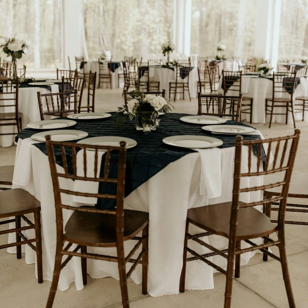 table set with white plates on white napkins on a black cloth over draped white tablecloth, surrounded by brown wooden chairs with chairs and table. There are several similar tables in the background table set with white plates on white napkins on a black cloth over draped white tablecloth, surrounded by brown wooden chairs with chairs and table. There are several similar tables in the background