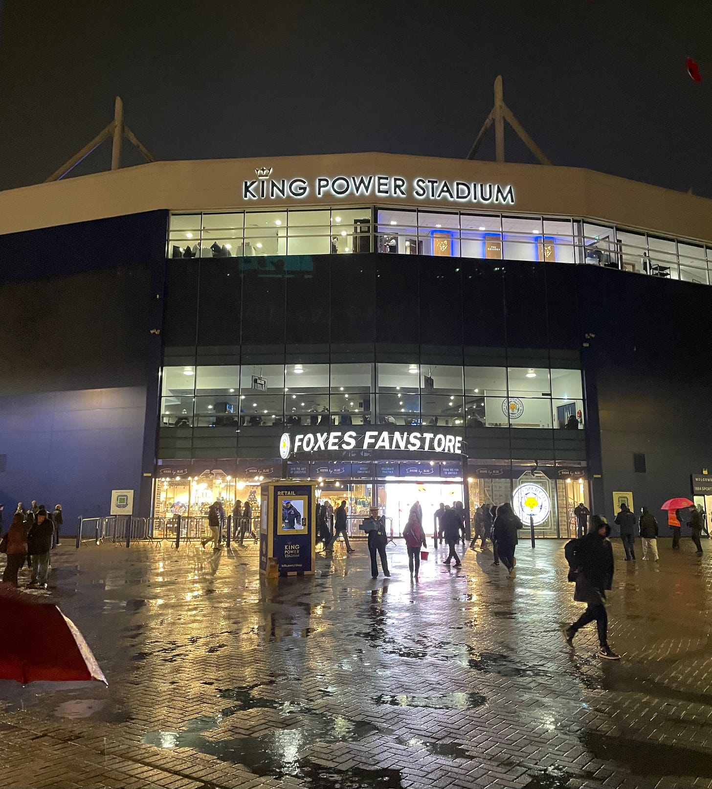 The front of the King Power Stadium in the rain, with the lights reflecting on the wet pavement.