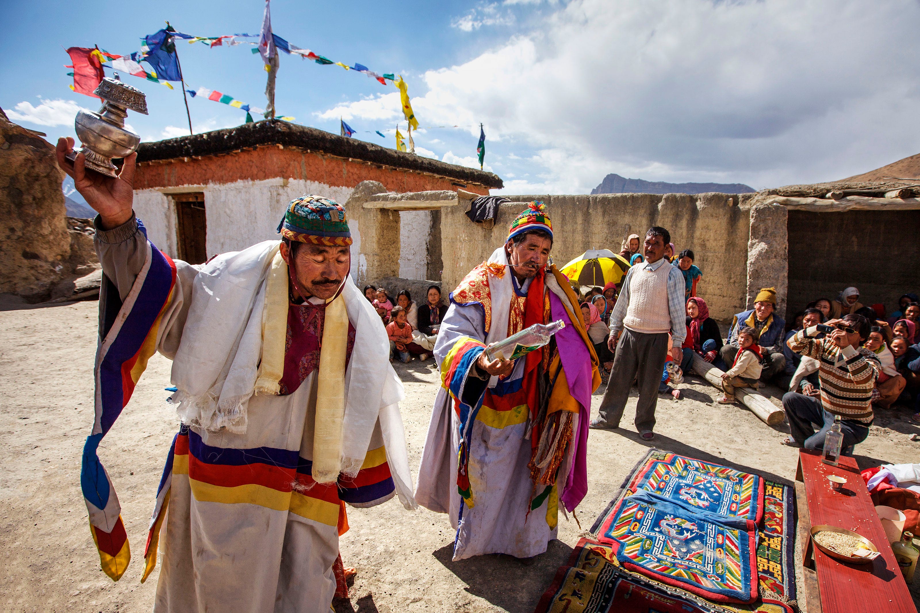 Two people in colorful traditional attire perform a ritual outdoors, holding objects, while a seated crowd watches. Prayer flags and a painted cloth are visible in the vibrant, sunlit scene.