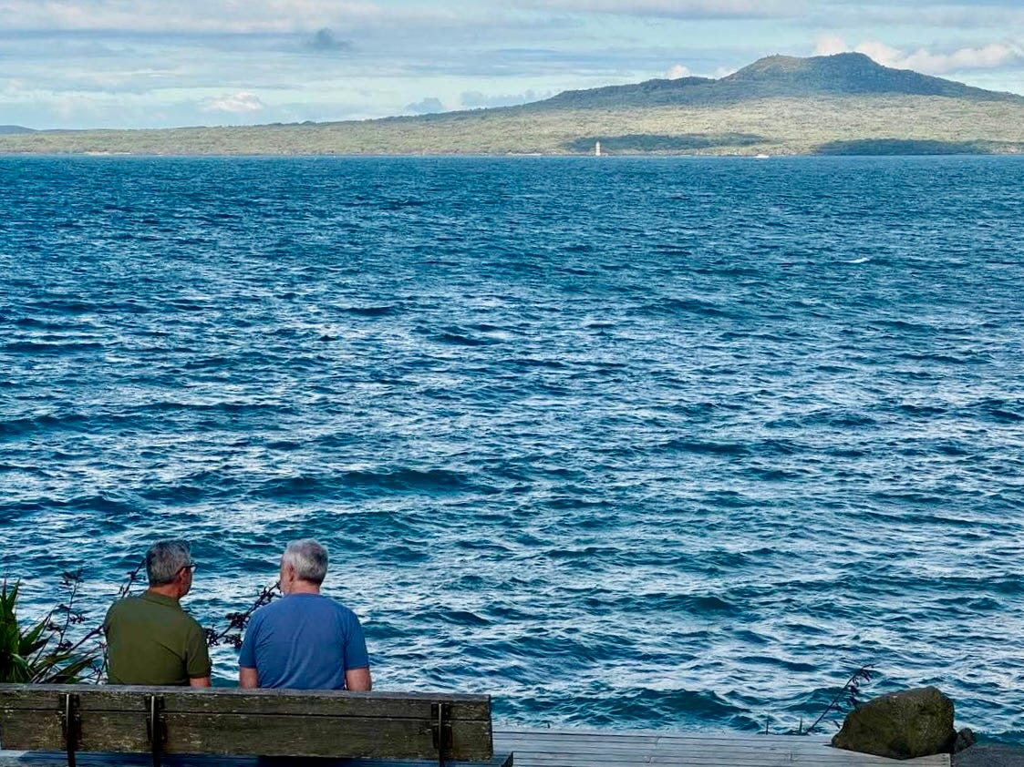 Two older men sit on a wooden bench facing the ocean, engaged in conversation. The sea stretches out before them with small waves rippling across its surface. In the background, a lush green island with a volcanic peak rises against a partly cloudy sky. The setting appears peaceful and reflective.