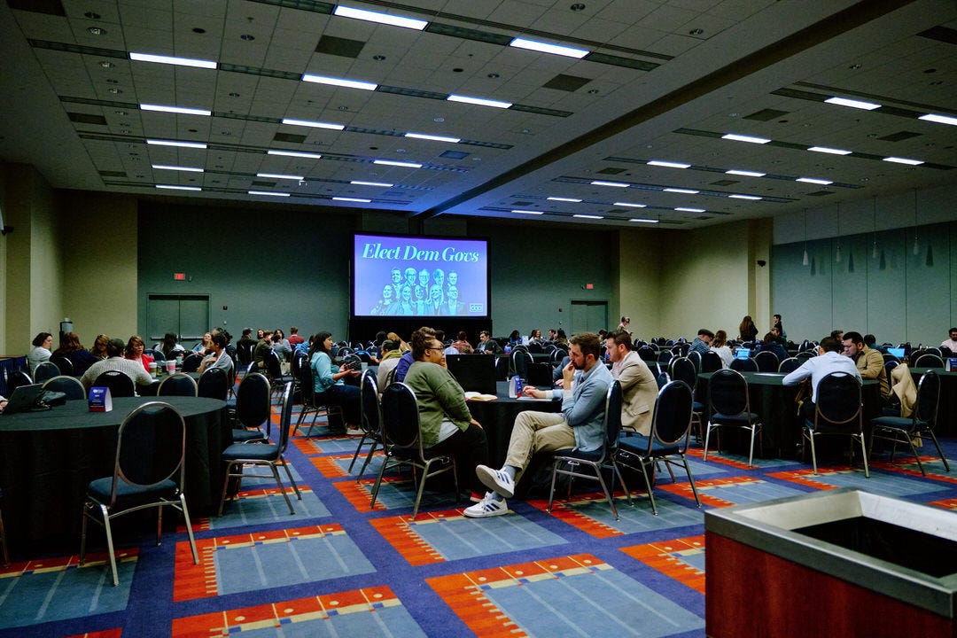 Large conference ballroom with scattered attendees at round tables during lunch, a projection screen at the front reading Elect Dem Govs in white text on blue