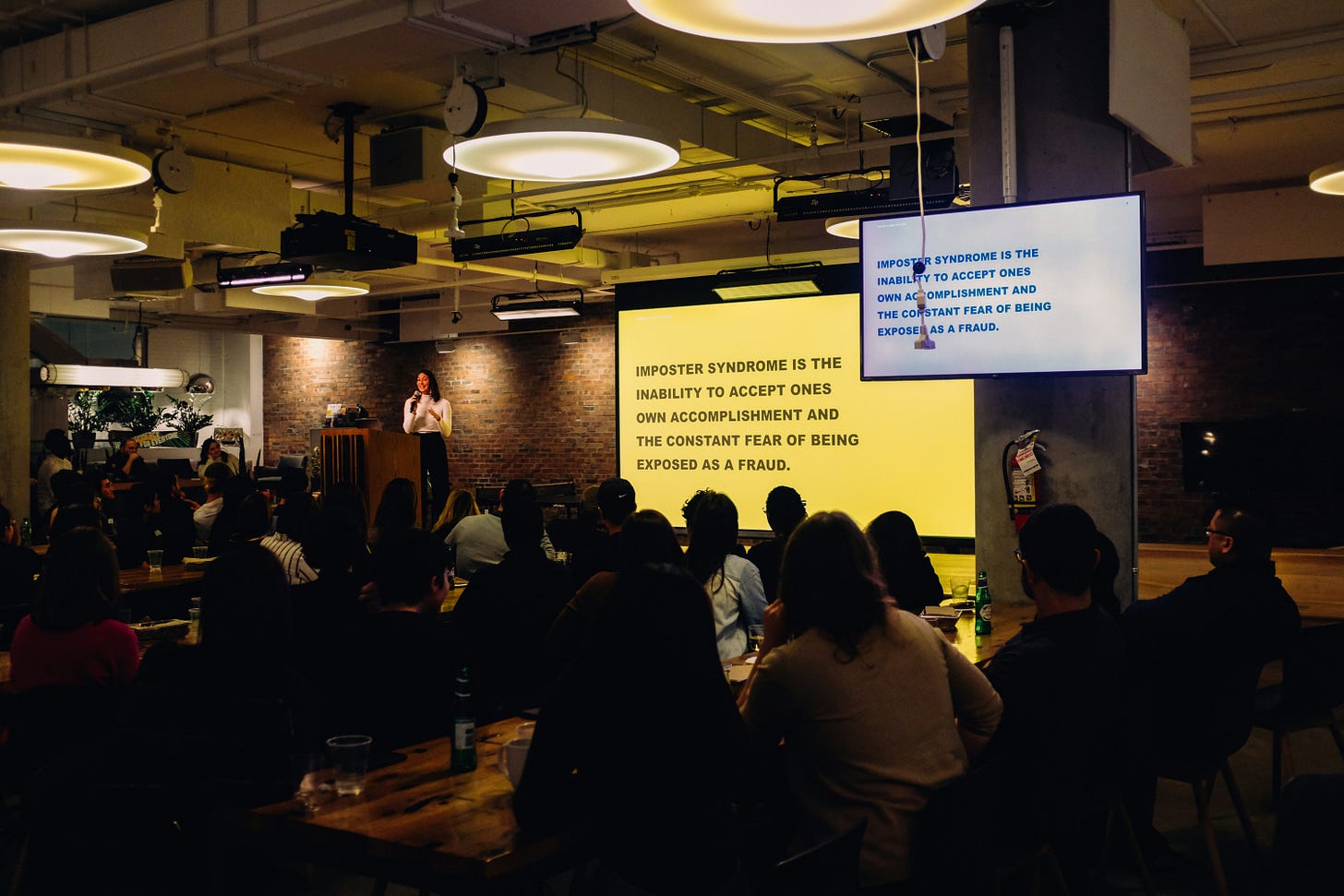 Woman presenting in front of an audience. A large projected slide states "Imposter syndrome is the inability to accept ones own accomplishment and the constant fear of being exposed as a fraud." Woman presenting in front of an audience. A large projected slide states "Imposter syndrome is the inability to accept ones own accomplishment and the constant fear of being exposed as a fraud."