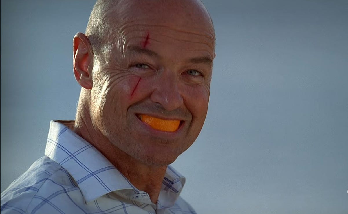 a scene from the tv show Lost: a bald man with a fresh cut over his eye smiles at the camera with a mouth full of orange peel