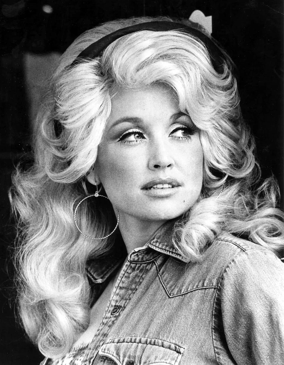 Parton from 1977 black and white publicity photo headshot; wearing a denim shirt, head turned to side, blonde hair everywhere