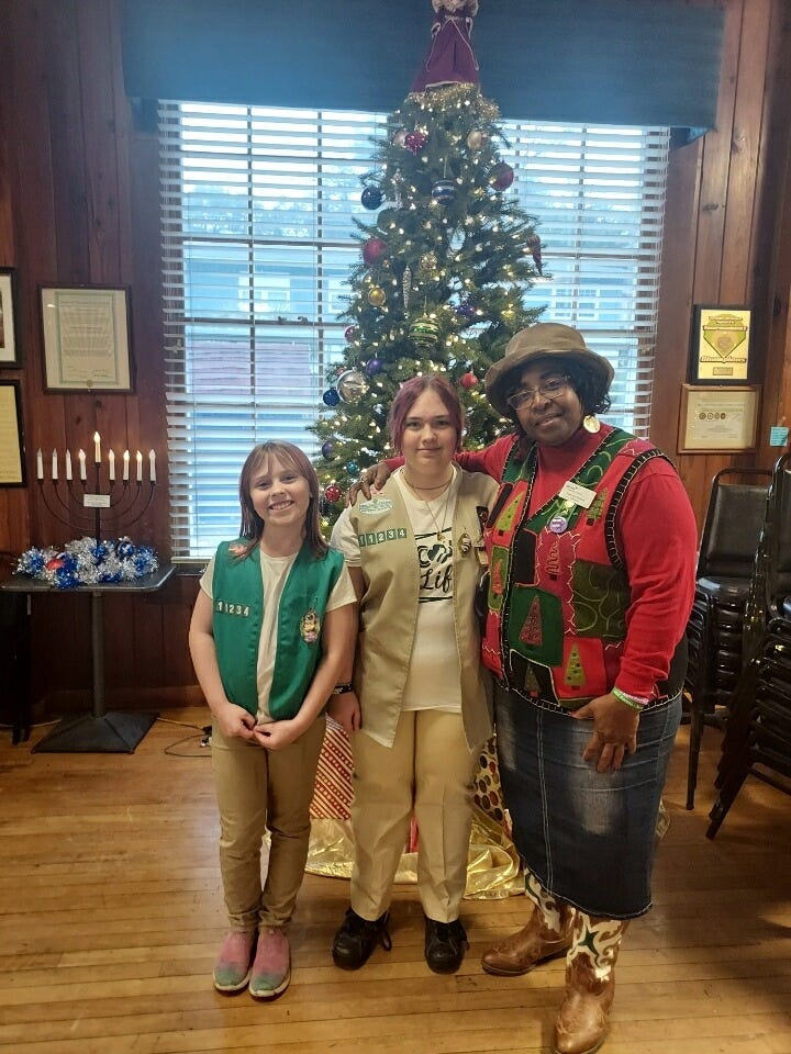 A festive indoor scene with a decorated Christmas tree topped by an angel and surrounded by wrapped presents. In front stand a bearded man in a green “U.S. ARMY” T‑shirt and two girls wearing scout uniforms—one in a tan vest with badges holding small items, the other in a green vest—next to several large filled bags. The room has wood-paneled walls, a large window with blinds behind the tree, framed certificates, a nine‑candle candelabrum resembling a menorah on the left, and stacked chairs on the right. Also, Three people pose indoors in front of a decorated Christmas tree: two youths wearing Girl Scouts uniform vests (one green, one tan) and an adult in a festive holiday vest, hat, denim skirt, and cowboy boots. The room has wood-paneled walls and a large window with blinds behind the tree. To the left is a table with a menorah and blue-and-white tinsel, while framed certificates hang on the walls and stacked chairs sit to the right on a hardwood floor. 
