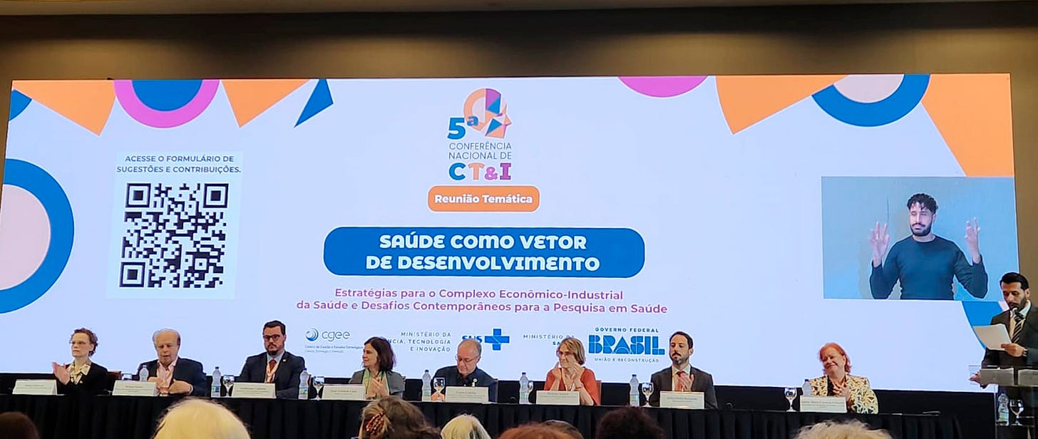 Em um palco, um homem em pé em um púlpito e quatro homens e quatro mulheres sentados à mesa. Atrás deles, uma grande tela digital, na cor branca, com figuras geométricas nas cores laranja, azul e rosa. Ao centro da tela, o texto 5ª Conferência Nacional de CT&I, Reunião Temática, Saúde Como Vetor de Desenvolvimento, Estratégias para o Complexo Econômico-Industrial da Saúde e Desafios Contemporâneos para Pesquisa em Saúde. De um lado do texto, um QR code, do outro lado, dentro de um quadro, um homem negro sinalizando em Libras e abaixo, as logos do Centro de Gestão e Estudos Estratégicos, do Ministério da Ciência, Tecnologia e Inovação, do SUS, do Ministério da Saúde e do Governo Federal.