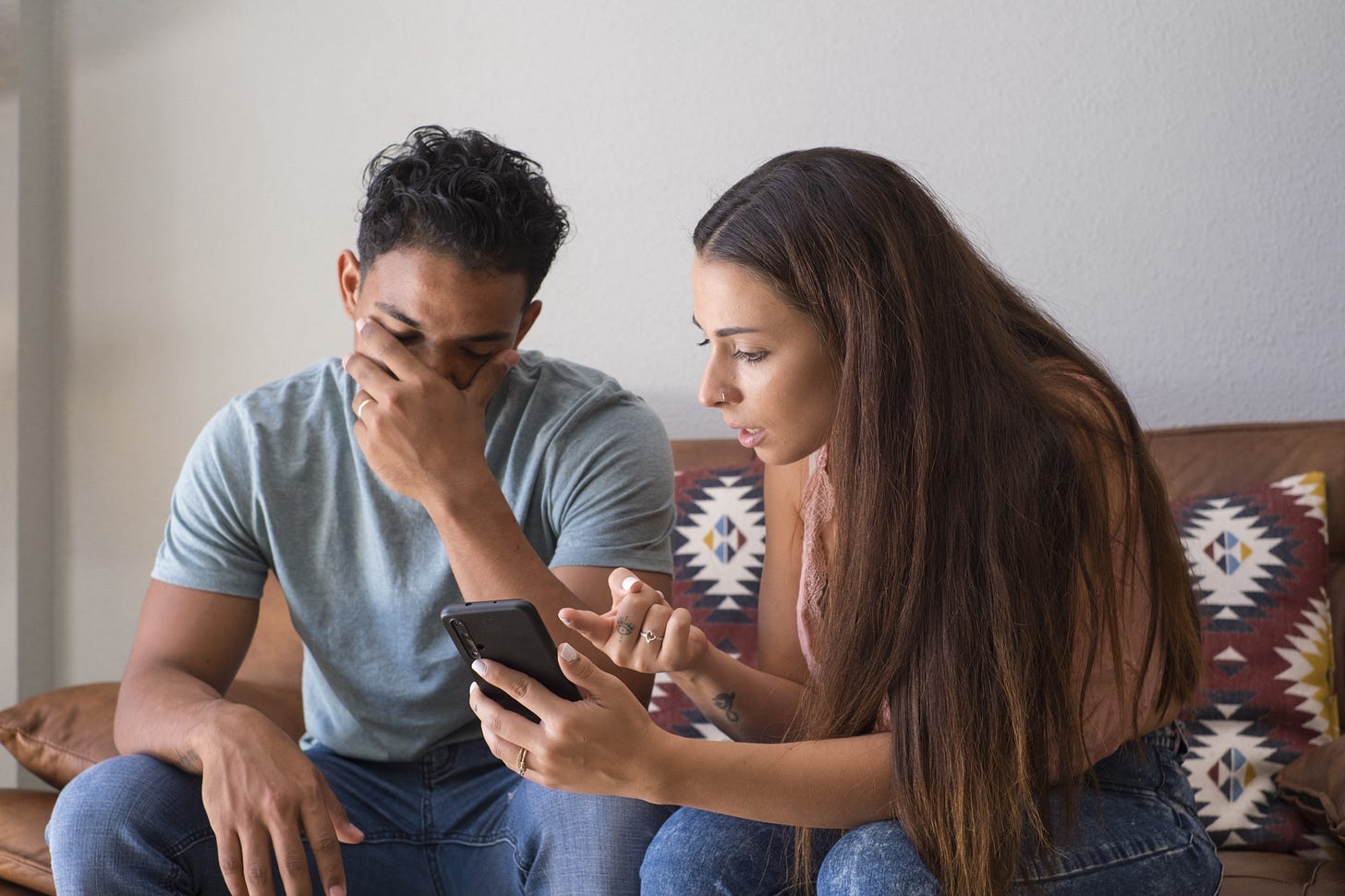 A man cradles his head in his hand while his partner jealousy rages over his phone.