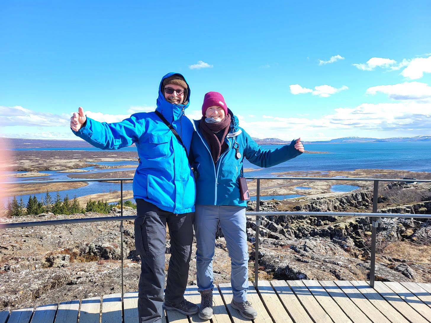 Man and woman wearing thick coats and hats smile, hands outstretched, side by side, on elevated ground overlooking land, distant sea, with blue sky above Man and woman wearing thick coats and hats smile, hands outstretched, side by side, on elevated ground overlooking land, distant sea, with blue sky above