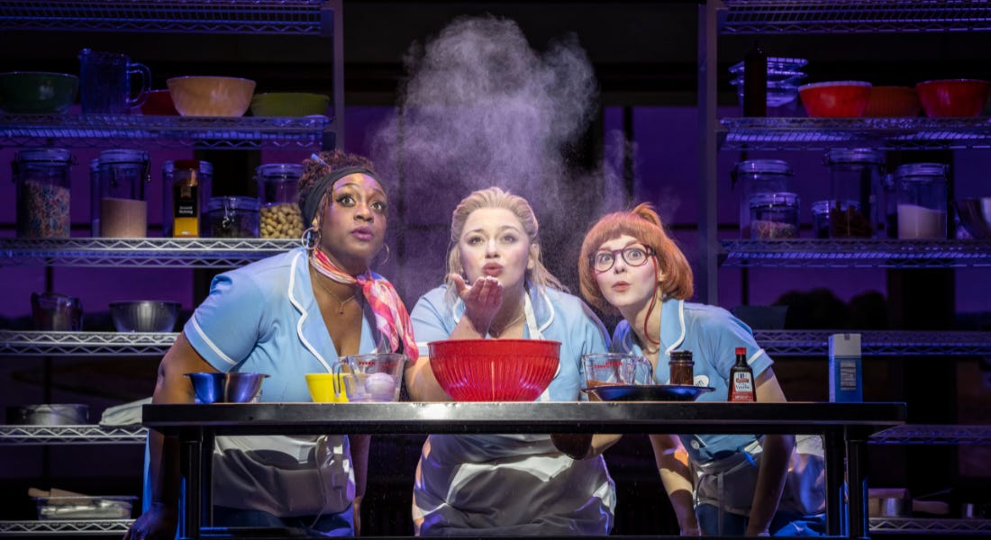 Three women in blue waitress uniforms on stage blowing flower, with ingredients behind them Three women in blue waitress uniforms on stage blowing flower, with ingredients behind them