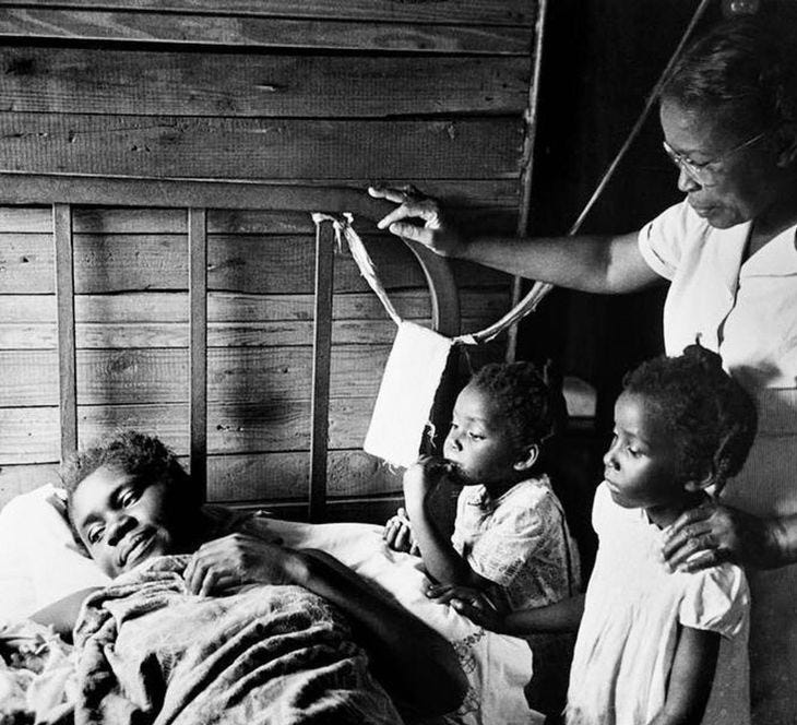 dark skinned black woman laying in bed, two small black girls beside her, and a Black woman behind them with a caring countenance.